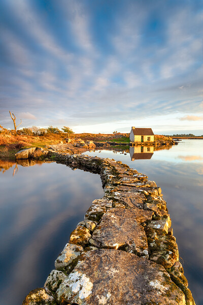 An old stone jetty across a lake leading to a small cottage Picture Board by Helen Hotson
