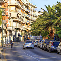 Buy canvas prints of Street landscape of the summer city of Loutraki, Greece, with passing cars and a teenager on a bicycle. by Sergii Petruk