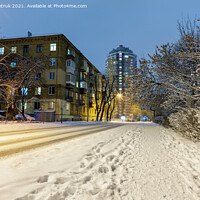Buy canvas prints of Snow-covered road along a city street with trees in the snow and city evening lighting against the backdrop of blue twilight. by Sergii Petruk