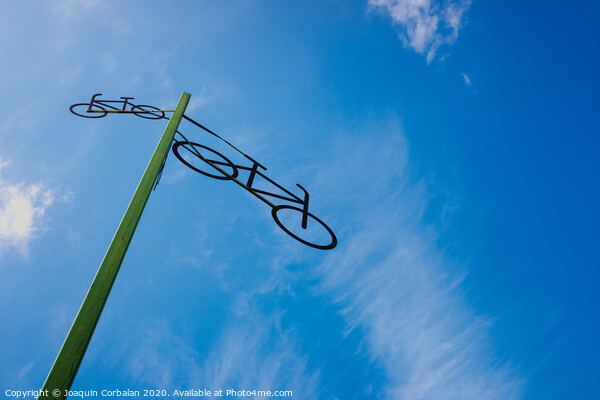 Post with the figure of some bicycles indicating the road, with blue sky and clouds in the background. Picture Board by Joaquin Corbalan