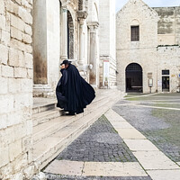 Buy canvas prints of Bari, Italy - March 12, 2019: A Catholic priest walks towards the cathedral dressed in a black cape and hat a day of wind and rain. by Joaquin Corbalan