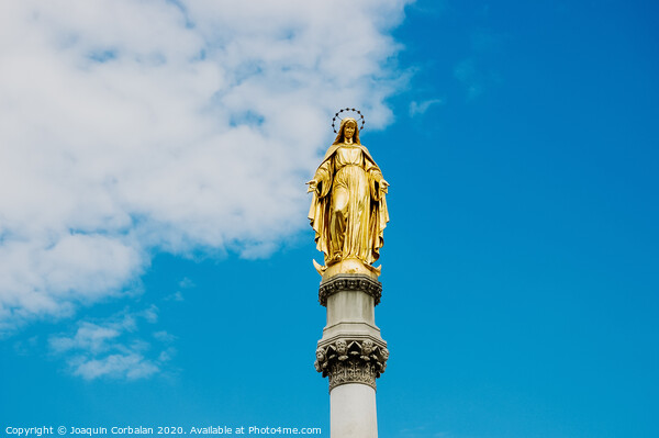 Golden religious statue, illuminated by the sun, of the Virgin Mary on top of a pedestal, with a background of blue sky and clouds. Picture Board by Joaquin Corbalan