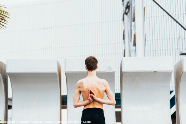 Young male athlete performing stretching exercises for back and arms in an urban place. Picture Board by Joaquin Corbalan