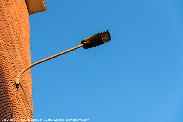 Lamppost on the red wall of a building, by day, with a blue sky background. Picture Board by Joaquin Corbalan