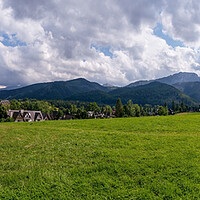 Buy canvas prints of Panoramic view of green grass meadow countryside field with A shaped houses against sleeping knight tatra mountain aka as giewont and dramatic clouds located in Zakopane, South Poland, Europe by Arpan Bhatia