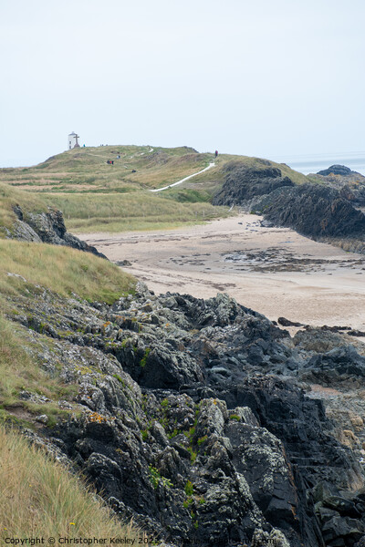 Rocky beach and lighthouse of Ynys Llanddwyn Picture Board by Christopher Keeley