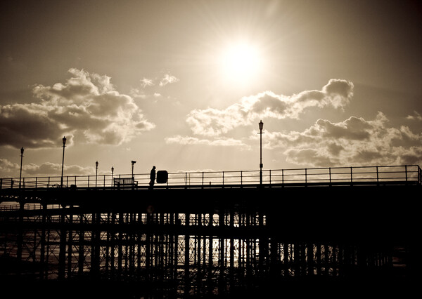 Sepia image of part of Southend Pier shot against the sun, with silhouette of structure and a man on the walkway. Southrnd on Sea, Essex. Picture Board by Peter Bolton