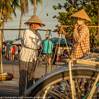 Buy canvas prints of Two Vietnamese women with conical hats, carrying shoulder poles by a river in the afternoon by SnapT Photography