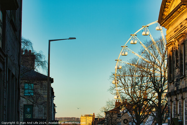 Urban sunset scene with silhouette of a Ferris wheel against a clear sky, flanked by historic buildings and a street lamp in Lancaster. Picture Board by Man And Life