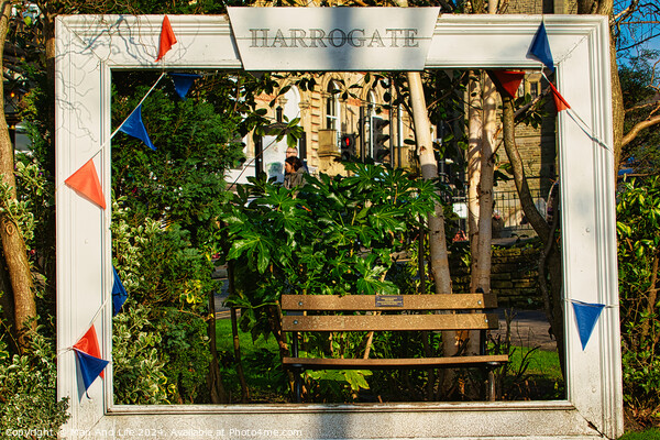 Quaint garden scene framed by a white wooden structure with 'Harrogate' sign, featuring a bench and lush greenery, adorned with colorful pennants. Picture Board by Man And Life