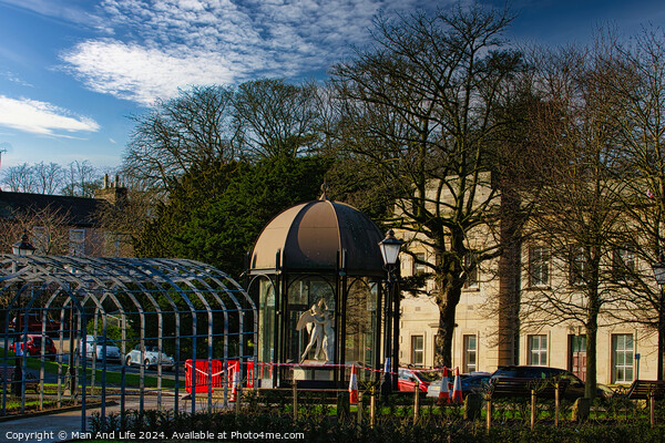 Urban park scene with modern glass pavilion, traditional street lamp, and lush trees under a blue sky with wispy clouds in Harrogate, England. Picture Board by Man And Life