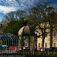 Buy canvas prints of Urban park scene with modern glass pavilion, traditional street lamp, and lush trees under a blue sky with wispy clouds in Harrogate, England. by Man And Life