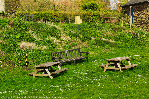 Rustic wooden benches and a table on a lush green grassy hillside, with a stone building and vegetation in the background, depicting a serene outdoor setting in York, North Yorkshire, England. Picture Board by Man And Life