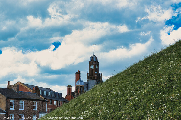 Quaint European town with historic buildings and a clock tower, set against a vibrant blue sky with fluffy clouds, and a lush green hill in the foreground in York, North Yorkshire, England. Picture Board by Man And Life