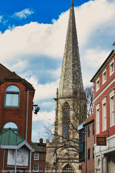 Vertical shot of an ancient church spire reaching into a blue sky with clouds, flanked by traditional brick buildings, showcasing architectural contrast and historical cityscape in York, North Yorkshire, England. Picture Board by Man And Life