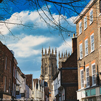 Buy canvas prints of Historic European city street with traditional brick buildings and a prominent Gothic cathedral in the background under a blue sky with clouds in York, North Yorkshire, England. by Man And Life