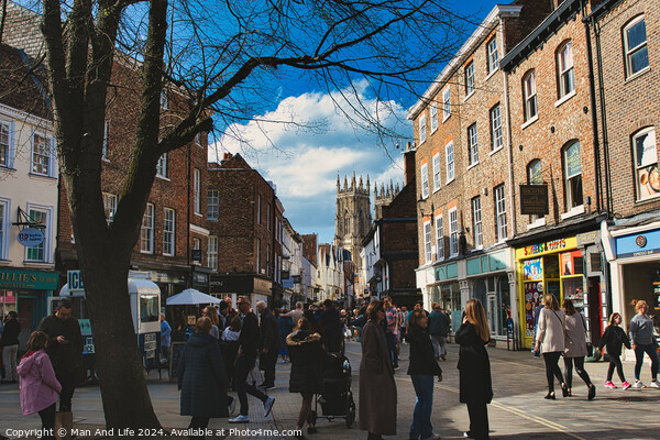 Bustling city street scene with pedestrians, historic buildings, and a cathedral spire under a blue sky with scattered clouds in York, North Yorkshire, England. Picture Board by Man And Life