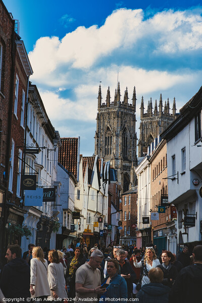 Bustling street scene with pedestrians in a historic city center, featuring old buildings and a prominent Gothic cathedral under a cloudy sky in York, North Yorkshire, England. Picture Board by Man And Life
