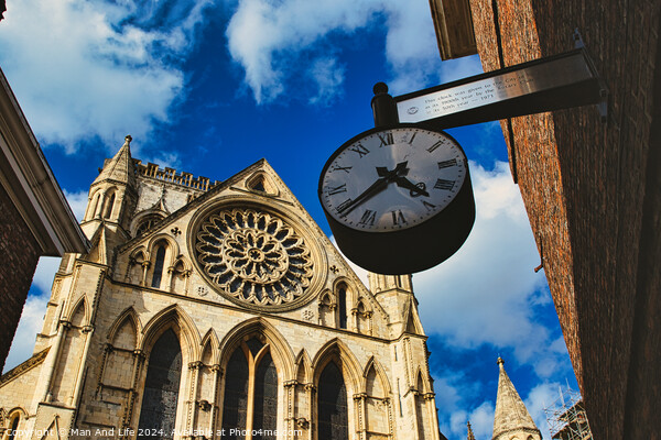Vintage street clock hanging with a gothic cathedral facade in the background, showcasing intricate architecture and a clear blue sky in York, North Yorkshire, England. Picture Board by Man And Life