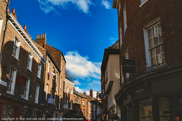 Charming European street scene with historic brick buildings under a clear blue sky with fluffy clouds, showcasing architectural details and local businesses in York, North Yorkshire, England. Picture Board by Man And Life