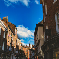 Buy canvas prints of Charming European street scene with historic brick buildings under a clear blue sky with fluffy clouds, showcasing architectural details and local businesses in York, North Yorkshire, England. by Man And Life