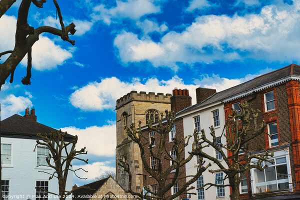 Quaint urban scene with historic stone tower, traditional buildings, and bare tree branches against a vibrant blue sky with fluffy white clouds in York, North Yorkshire, England. Picture Board by Man And Life