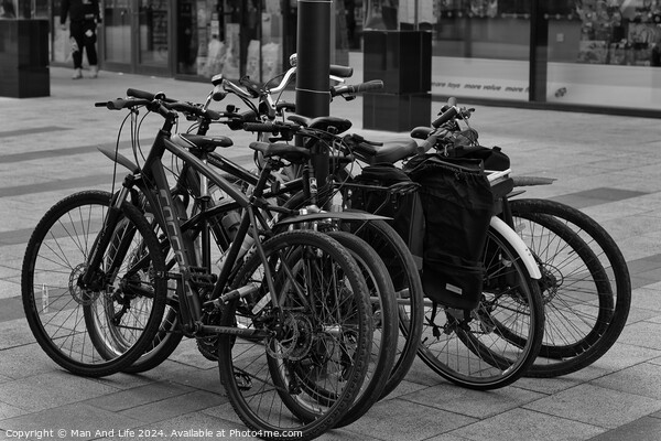 Black and white image of multiple bicycles locked to a bike rack in an urban setting, with a blurred background of a city street Picture Board by Man And Life